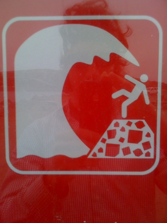 the reflection of a person who is taking a photo of a red sign that warns beach goers to beware of large waves